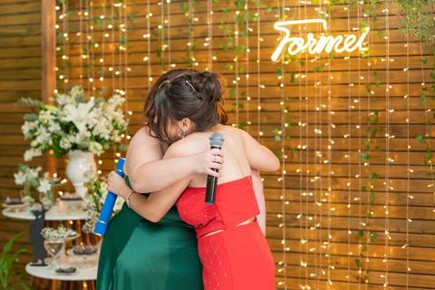 ALt text: a foto mostra as formandas se abraçando, uma usa vestido verde  longo e a outra um vestido vermelho longo e ambas tem cabelos castanhos'