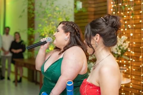 ALt text: a foto mostra uma formanda falando no microfone vestindo um vestido vermelho seus cabelos estão presos em um coque e são castanhos, enquanto a outra formanda sorri ela veste um vestido longo verde e também tem cabelos castanhos'