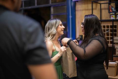 Alt text: uma mulher de cabelos e roupas pretas entrega uma sacola de presente para uma mulher loira de vestido verde'