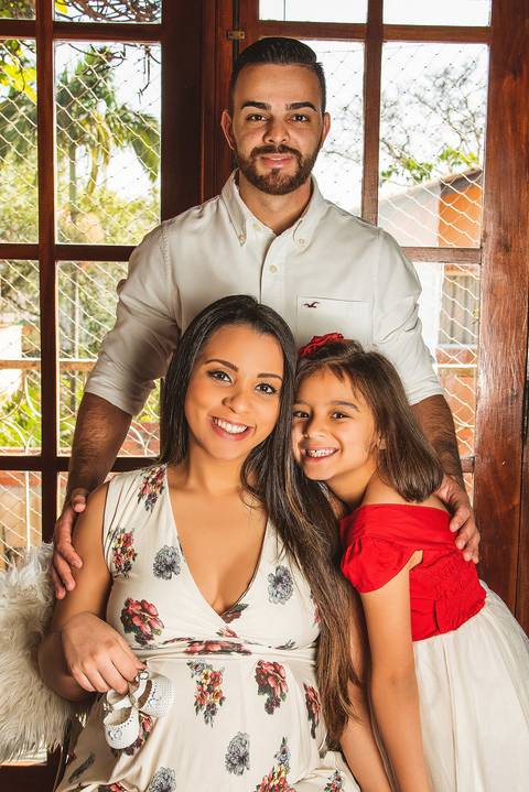 Foto da Mãe Sentada Com Sapatinho na Barriga Sendo Abraçada Pela Filha e o Marido em Pé Atras No Estúdio Jardim Guedala em São Paulo, Foto Feita Pela Priscila Felix Fotógrafa de Família, Casamento e Ensaios '