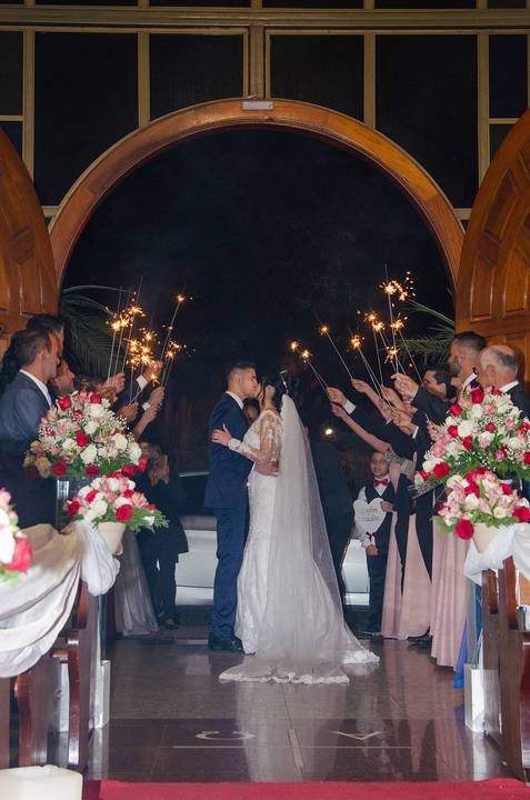 Fotografia dos Noivos Se Beijando Na Porta da Paróquia Nossa Senhora de Fátima e Em Volta os Padrinhos Com Velas Estrelas Acesas em Guarulhos SP Feita por Priscila Felix Fotógrafa de Casamentos, Ensaios e Família'
