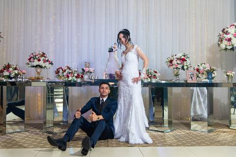 Fotografia dos Noivos Brincando em Frente à Mesa do Bolo Ela Em Pé Sorrindo e Ele Sentado no Chão Fazendo Joinha No Buffet Europa em Guarulhos SP Feita por Priscila Felix Fotógrafa de Casamentos, Ensaios e Família'