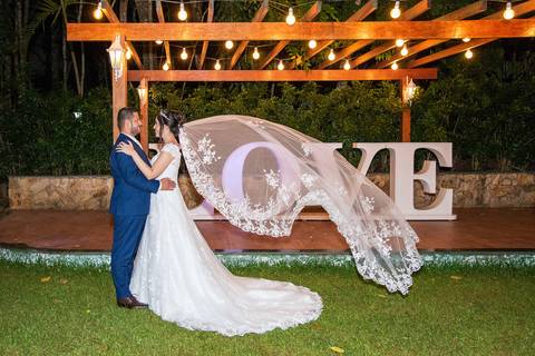 Fotografia dos Noivos Na Área Externa Em Frente Ao Deck Iluminado O Véu da Noiva Voando Em Frente Da Palavre 