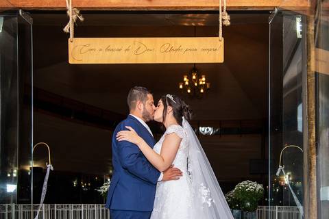 Fotografia dos Noivos Se Beijando Na Entrada do  Salão de Cerimônia do Casamento Embaixo De Uma Placa Dizendo 
