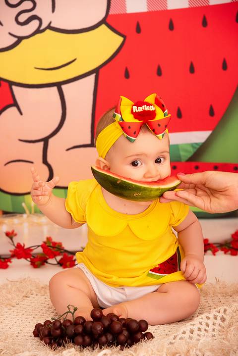 Fotografia da Bebê Comendo Um Pedaço de Melancia da Mão da Mãe no Estúdio Com Cenário da Magali da Turma da Monica em SP Feita por Priscila Felix Fotógrafa de Casamentos, Ensaios e Família'