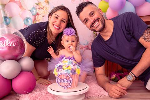 Fotografia da Bebê Sentadinha Entre os Pais Com o Dedo na Boca no Estúdio Com Cenário Rosa Com Bexigas Personalizadas em SP Feita por Priscila Felix Fotógrafa de Casamentos, Ensaios e Família'