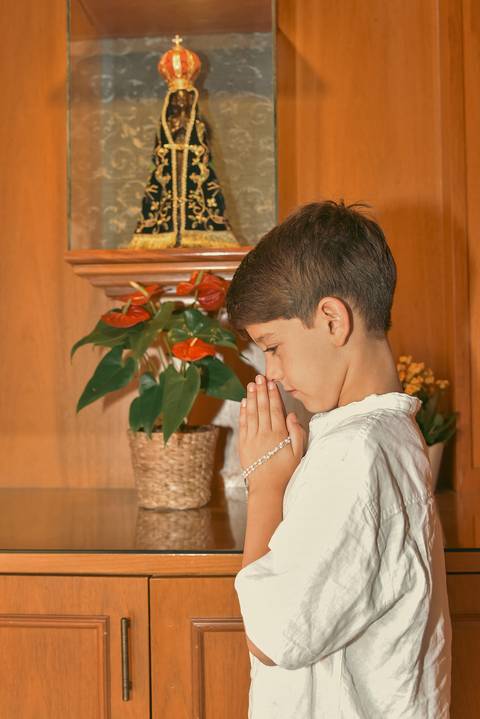 Fotografia do Batizado Segurando o Terço De Perfil Em Oração Em Frente a Imagem de Nossa Senhora Na Igreja Verbo Divino em São Paulo, SP - Feita por Priscila Felix Fotógrafa de Casamentos, Ensaios e Família'