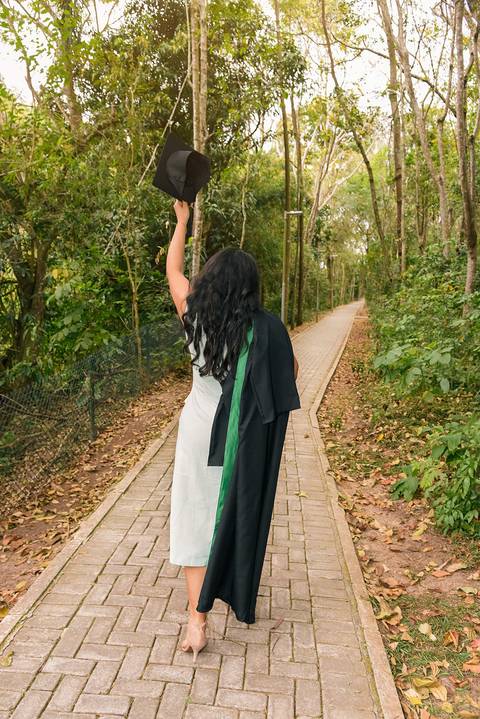 Foto da Formanda Caminhando de Costas Com a Beca Solta No Ombro e Na Esquerda Erguendo O Canudo No Parque do Pau D´Alho Em Guararema - SP, Foto Feita Pela Priscila Felix, Fotógrafa de Família, Casamento e Ensaios '