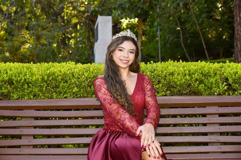 Foto da Debutante Sentada Em Frente Ao Jardim de Pernas Cruzadas, Mão Nos Joelhos Usando Um Vestido Vermelho Curto E Uma Coroa No Parque da Água Branca Em São Paulo - SP, Foto Feita Pela Priscila Felix Fotógrafa de Família, Casamentos e Ensaios '