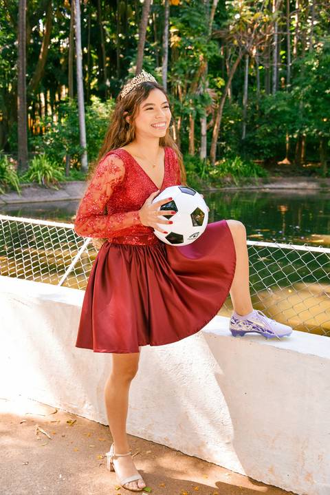Foto da Debutante Em Frente Ao Lago Usando o Vestido Vermelho, A Coroa, Uma Chuteira Lilás, Segurando Uma Bola De Futebol No Parque da Água Branca Em São Paulo - SP, Foto Feita Pela Priscila Felix Fotógrafa de Família, Casamentos e Ensaios '
