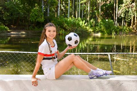 Foto da Debutante Em Frente Ao Lago Usando Um Shorts Branco, A Camisa do SP, Uma Chuteira Lilás, e Segurando Uma Bola De Futebol No Parque da Água Branca Em São Paulo - SP, Foto Feita Pela Priscila Felix Fotógrafa de Família, Casamentos e Ensaios '