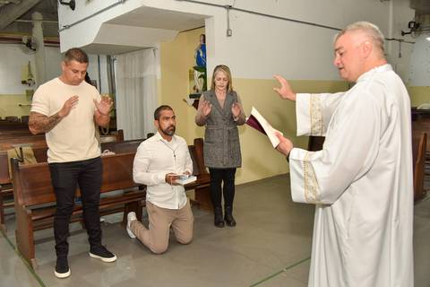 Fotografia Do Rafael De Joelhos, Após Receber a Medalha, O Livro De Orações E A Bíblia E O Padre Orando Com A Mão Em Sua Direção No Altar Na Paróquia São Judas Tadeu em São Paulo, SP Feita por Priscila Felix Fotógrafa de Casamentos, Ensaios e Família'