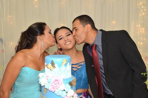 Fotografia da Debutante Com Seus Pais a Beijando Na Bochecha Na Mesa do Bolo Com Top Azul e Saia Longa Florida em Itaquera SP Feita por Priscila Felix Fotógrafa de Casamentos, Ensaios e Família'