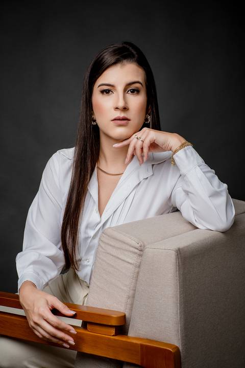 Retrato profissional feminino de mulher sentada em poltrona de madeira, vestindo camisa branca elegante, com a mão próxima ao rosto e fundo escuro dramático.'