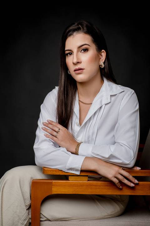 Advogada em postura relaxada e elegante, sentada em poltrona de madeira com camisa branca, braço apoiado, olhar direto e fundo preto.'