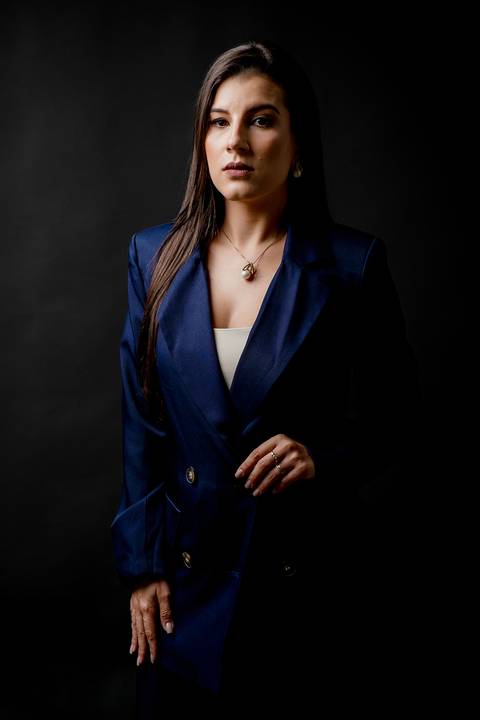 Retrato de autoridade com advogada em pé vestindo blazer azul marinho, iluminação contrastante contra fundo preto profundo.'