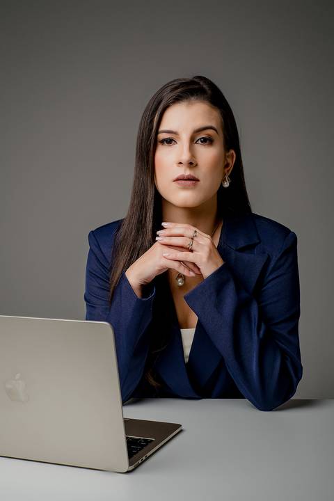 Advogada sentada à mesa com notebook prata, vestindo blazer azul marinho e colar de pérolas, olhando para a câmera com expressão séria contra fundo cinza.'
