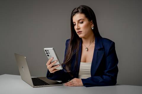 Profissional de terno azul marinho olhando para a tela de um smartphone prata, com um notebook aberto sobre a mesa clara, fundo cinza.'