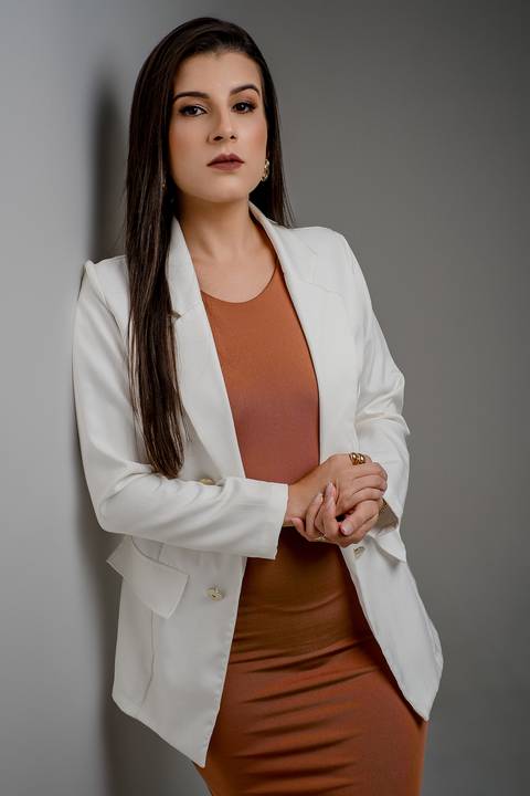 Executiva encostada em parede cinza, vestindo blazer branco estruturado sobre vestido terracota, com braços cruzados e postura confiante.'