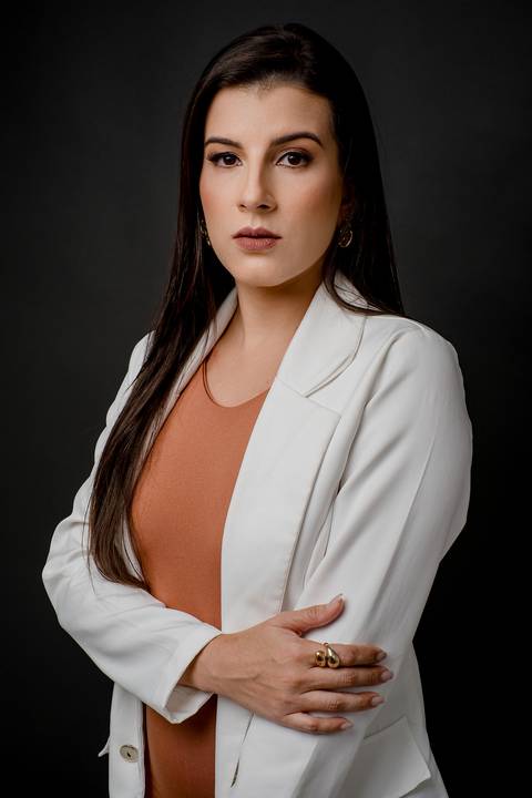 Fotografia de posicionamento com mulher vestindo blazer branco e vestido terracota, braços cruzados, olhar assertivo e fundo preto texturizado.'