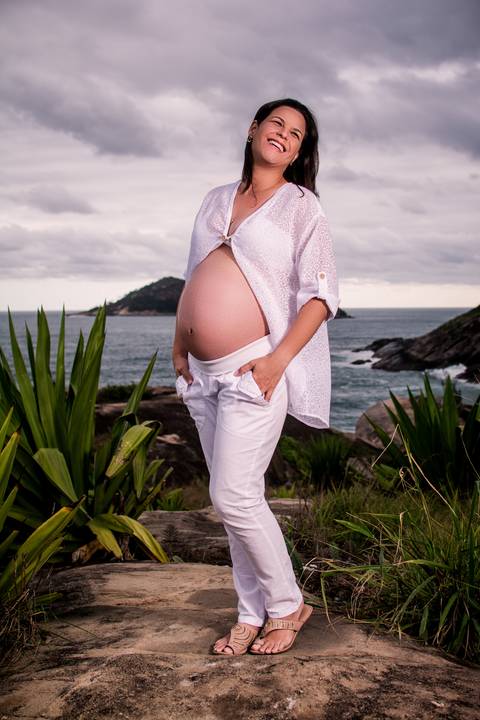 Ensaio fotográfico gestante realizado na Praia de Grumari, grávida vestindo calça e blusão blusão branco posando em pé com as mãos no bolso'