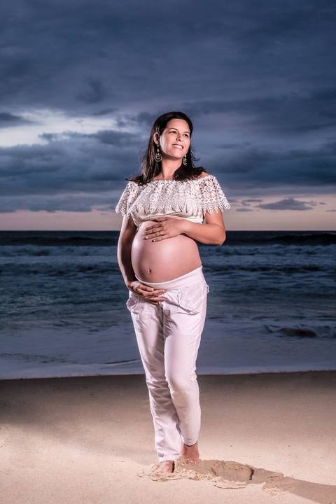 Ensaio fotográfico gestante na praia de Grumari, grávida vestida com calça branca e croped de renda off-white, segurando a barriga e sorrindo ao pôr-do-sol na praia'