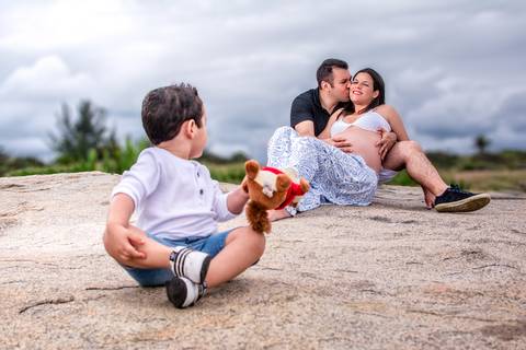 Suzana com marido e filho na praia de grumari para ensaio gestante, davi olhando para trás vendo o pai beijar a mamãe'