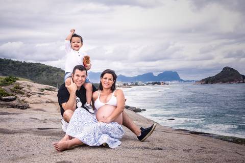ensaio gestante na praia de grumari, criança na carcunda do pai que está sentado na pedra abraçando a esposa grávida'