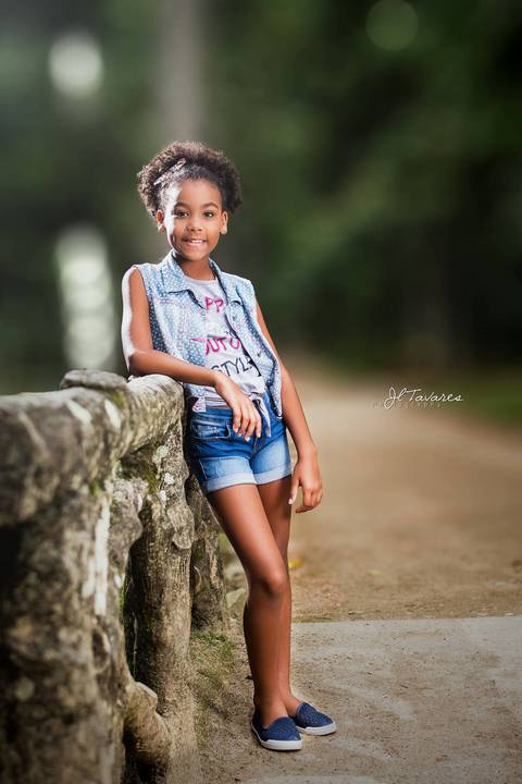 Milena Pinheiro Vestindo Shorts e camisa jeans com blusa branca por dentro encostada no corrimão de madeira durante o ensaio infantil no Jardim Botânico do Rio de Janeiro.'