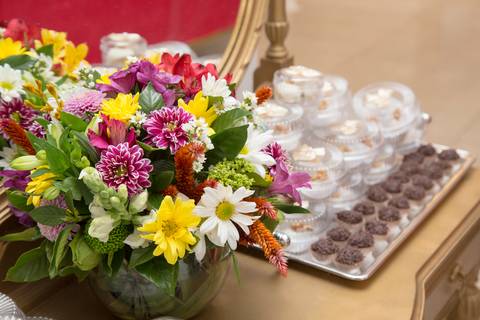 Arranjo floral exuberante em primeiro plano, com doces finos e coloridos levemente desfocados ao fundo, compondo a decoração da festa de 50 anos em São Paulo.'