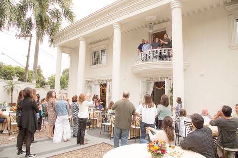 Aniversariante de 50 anos e amigos na sacada da mansão em São Paulo, com um plano aberto que inclui convidados na parte de baixo e a grandiosidade da mansão, capturando a amplitude da festa.'