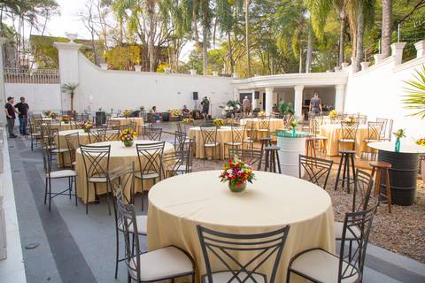 Mesas elegantemente decoradas com arranjos de flores na área externa de uma mansão em São Paulo, prontas para receber os convidados da festa de 50 anos, em meio a um paisagismo cuidado.'