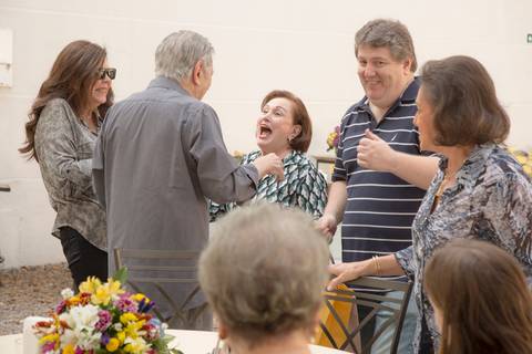A mãe do aniversariante e o próprio aniversariante de 50 anos rindo e interagindo com convidados, compartilhando a alegria da festa em São Paulo.'