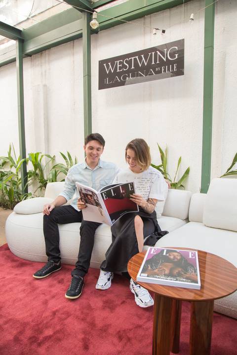 Duas participantes do evento Westwing + ELLE no Restaurante Chou em São Paulo, compartilhando o sofá modular e folheando a revista ELLE juntas, em um momento de leitura e interação.'