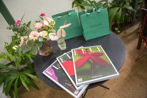 Mesa de boas-vindas no evento Westwing + ELLE no Restaurante Chou em São Paulo, exibindo revistas da ELLE, sacolas personalizadas da Westwing e um belo arranjo de flores, convidando à experiência.'