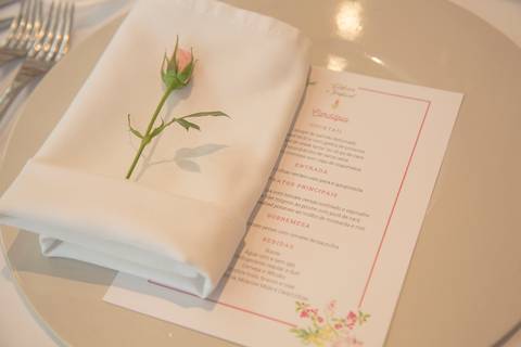 Detalhe de um menu de casamento personalizado sobre a mesa no Restaurante Cantaloup, com um guardanapo de tecido impecavelmente dobrado ao lado e uma flor delicada repousando sobre ele.'