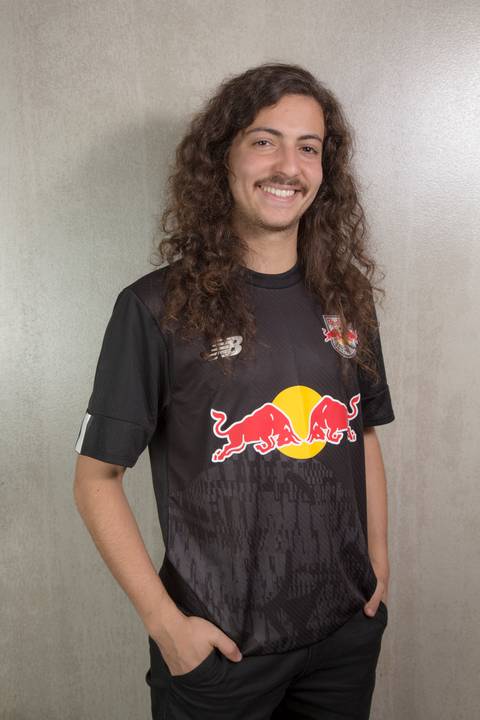 Retrato de um jovem estagiário vestindo uma camiseta com a marca Red Bull, sorrindo e olhando para a câmera, na sede da empresa em São Paulo.'