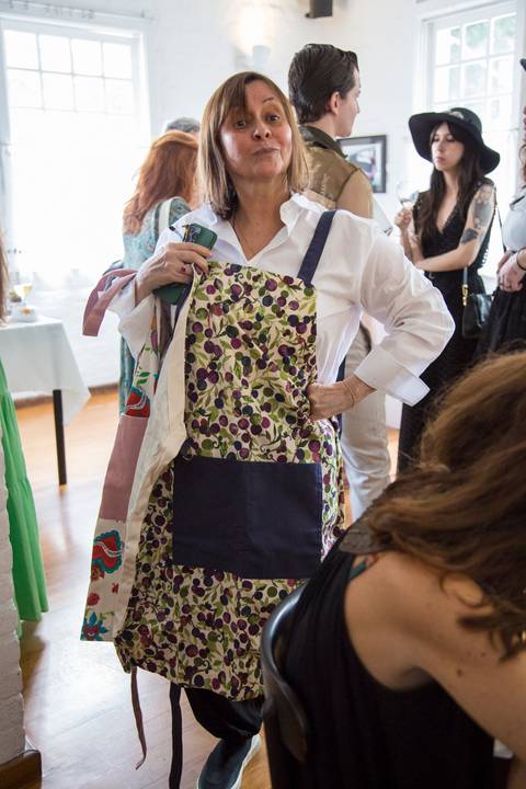 Chef Carla Pernambuco exibindo um avental de sua coleção em parceria com a Westwing, com um sorriso, durante o evento de lançamento no Restaurante Carlota em São Paulo.'