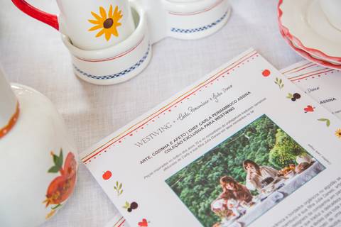 Papel informativo sobre a parceria Westwing com Carla Pernambuco e Julia Danesi, posicionado sobre uma mesa e cercado pelas louças da coleção, no evento do Restaurante Carlota em São Paulo.'