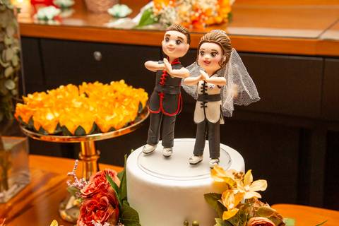Topo do bolo de casamento com bonequinhos personalizados dos noivos fazendo a pose de Kinrai do kung fu, vestidos com o uniforme da escola Kwan Kun, celebrando a paixão do casal no Restaurante Le Bife em São Paulo.'