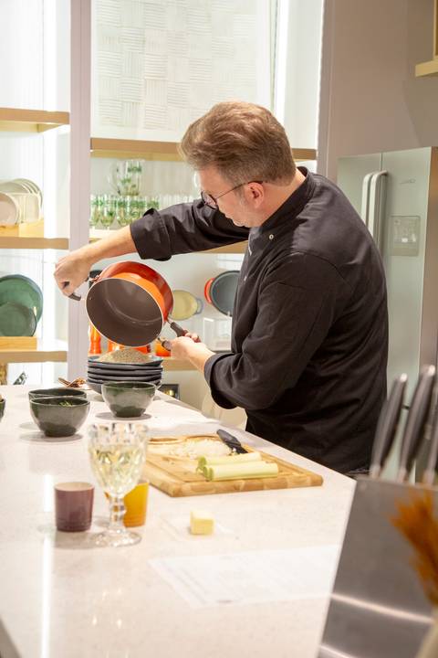 Chef Carlos Bertolazzi cozinhando ativamente o risoto, demonstrando técnicas culinárias no workshop do evento Westwing + Porto Brasil na loja de Moema, São Paulo.'