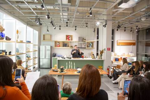 Participantes do workshop de risoto em primeiro plano, com o Chef Carlos Bertolazzi ao fundo da imagem, no palco da loja Westwing de Moema, São Paulo, demonstrando o foco da audiência.'