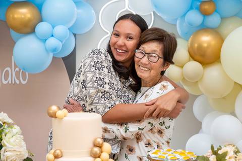 A aniversariante japonesa de 80 anos recebendo um abraço emocionante de sua neta ao lado da mesa do bolo, um gesto de carinho na festa em Boituva-SP.'