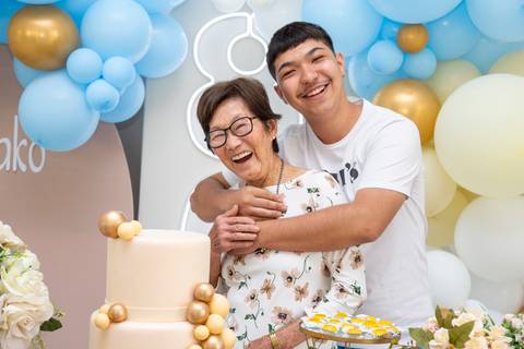 A aniversariante japonesa de 80 anos sendo abraçada por seu neto na mesa do bolo, expressando o afeto entre avó e neto na celebração em Boituva-SP.'