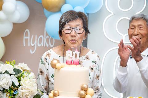 A aniversariante japonesa de 80 anos soprando as velas de seu bolo de aniversário com um sorriso, realizando um desejo na celebração em Boituva-SP.'