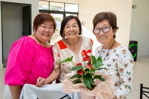 A aniversariante japonesa de 80 anos segurando uma flor, posando com duas convidadas, em um momento de carinho e gratidão na festa em Boituva-SP'