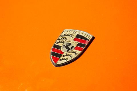 Emblema clássico do Porsche em close-up, com um vibrante fundo laranja que complementa o icônico design da marca, na oficina DAN911 em São Paulo.'