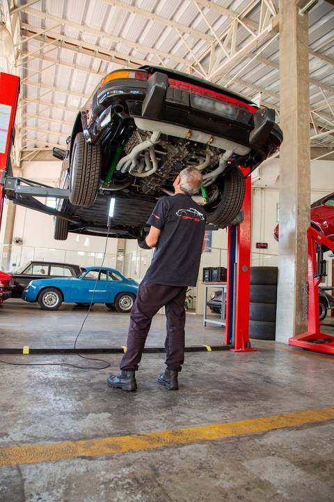 ecânico da DAN911 em São Paulo avaliando a parte inferior de um Porsche suspenso no elevador, com foco na inspeção técnica detalhada do chassi e componentes.'