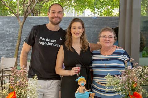 Futuros pais sorrindo e posando para foto com uma convidada durante o chá de bebê em São Paulo, registrando a presença de amigos e familiares.'