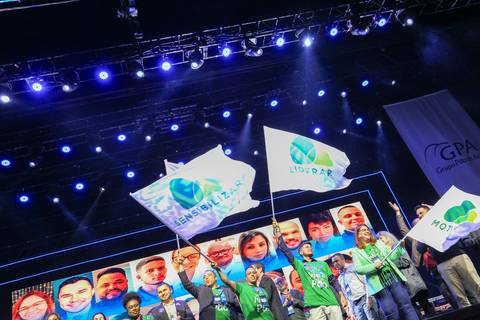 Um grupo de convidados do GPA balançando vigorosamente as bandeiras da empresa no palco do Espaço Unimed em São Paulo, em um momento de entusiasmo e celebração.'